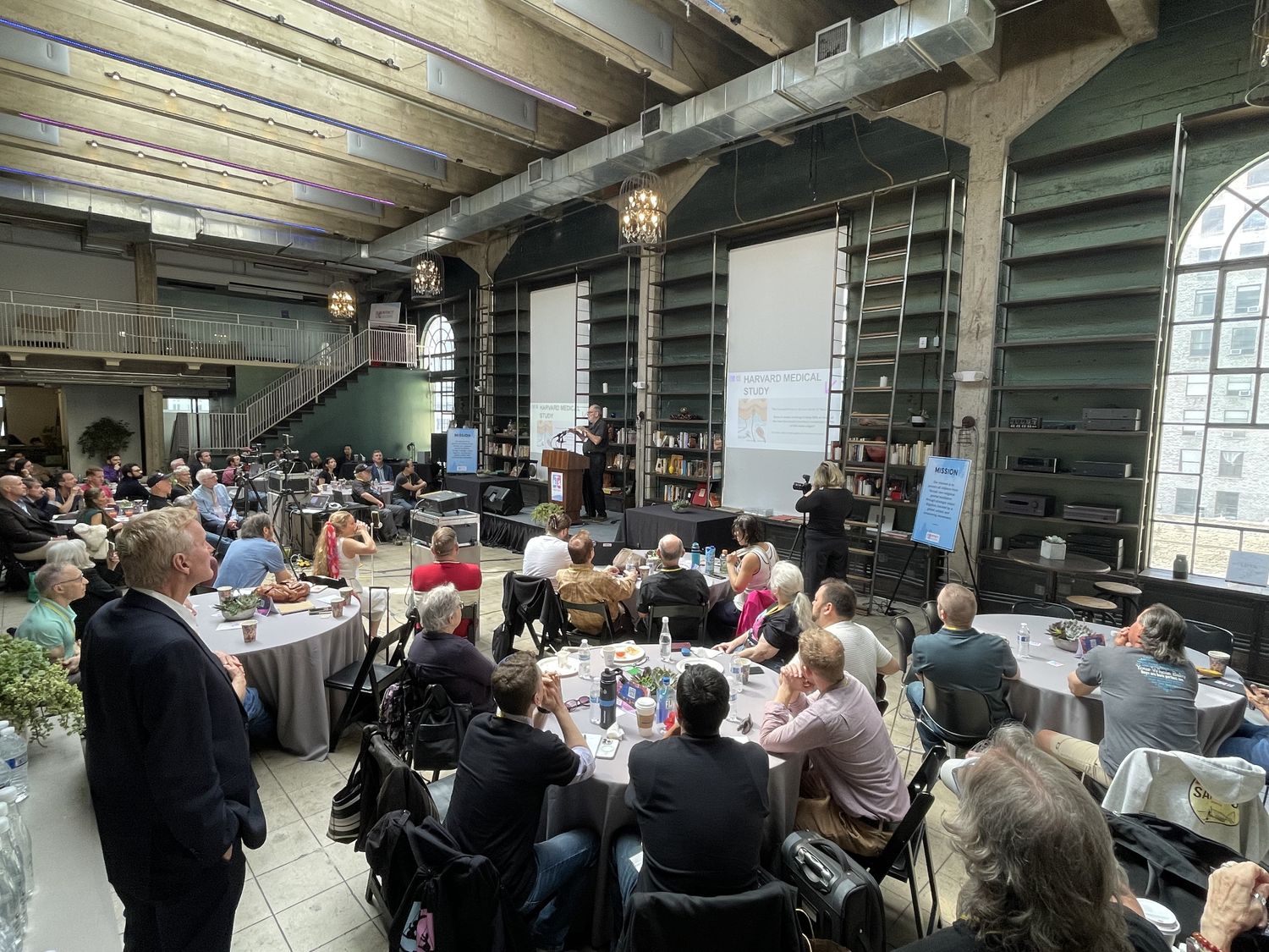 Conference audience seated at round tables inside Historic Dancehall, viewed from the bar